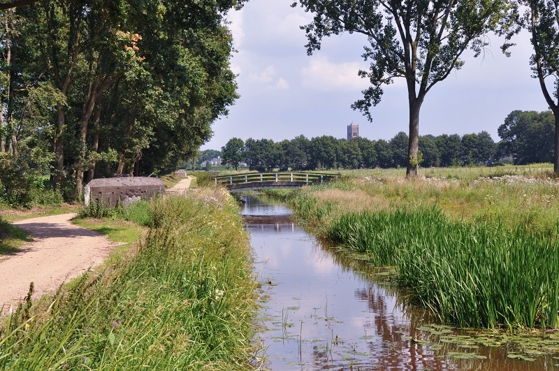 Afbeelding van een gedeelte van de Verborgen Raamvallei. Naast een wandelpad met oude bunker ligt De Raam, de beek die door dit gebied stroomt. Over de Raam ligt een brug naar een bloemrijk weiland. In de verte is de kerktoren van Mill te zien, achter een